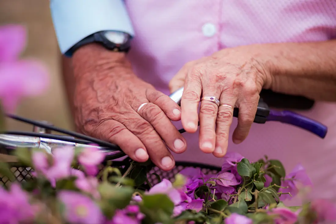 Una pareja de personas mayores se toma de las manos junto a unas flores